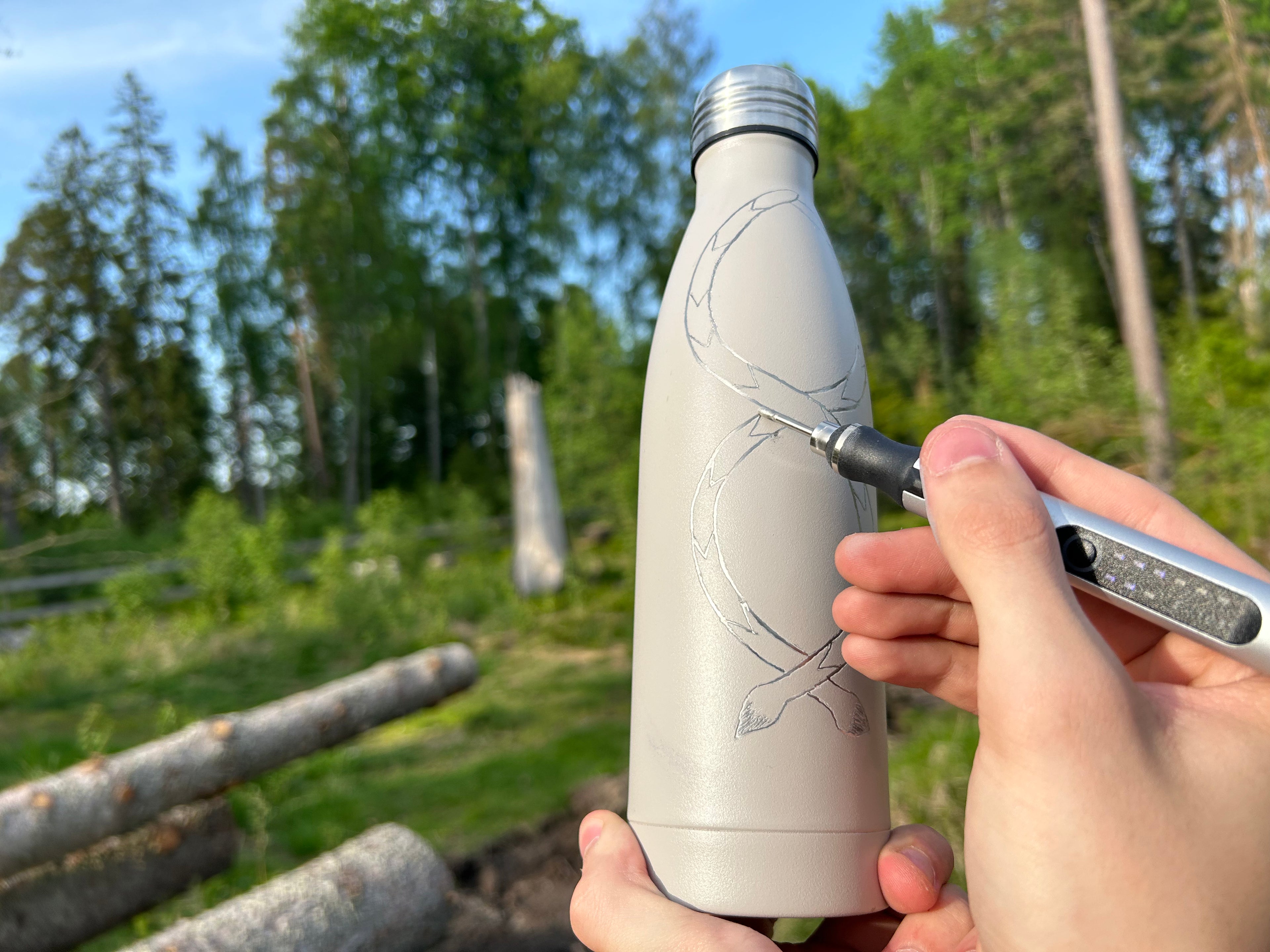 Ormmönster på en metallflaska. Skogen syns i bakgrunden och graveringspennan hålls mot flaska. 